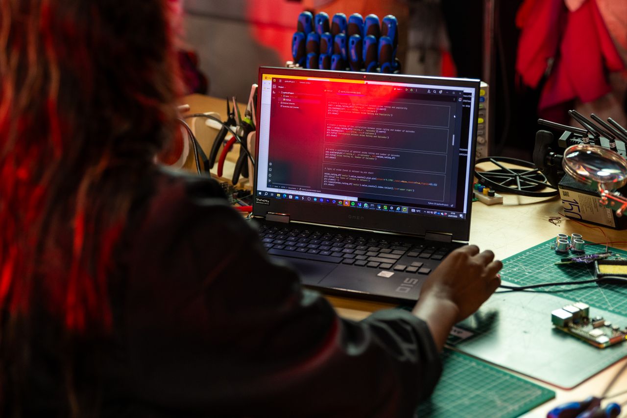 Home A software developer writing code on a laptop at a cluttered workbench with electronic tools.